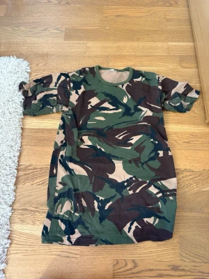 Camouflage t-shirt i bomull - Säljer en klassisk t-shirt med camouflage-mönster i grönt, beige och brunt. T-shirten har rund halsringning och korta ärmar. Perfekt för dig som gillar streetwear eller vill sticka ut lite extra.