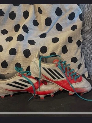 Vita och röda fotbollsskor med blå snören - Snygga fotbollsskor i vitt och rött med blå snören och svarta ränder på sidan. Skorna är väldigt sällsynta från f50 serien som användes mellan 2011-2015. Skorna har klassisk låg profil och dobbar för spel på gräs. Tillverkade i syntetmaterial för bra hållbarhet och komfort på planen.