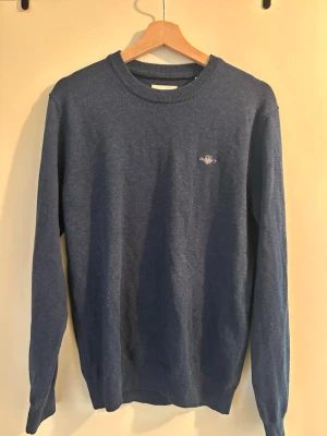 Blå GANT crewneck - Mysig blå stickad GANT crewneck med rund halsringning och diskret broderad logga på bröstet. Tröjan har långa ärmar och ribbade muddar vid ärmslut och nederkant. Perfekt för lager på lager och enkel att matcha till olika outfits.