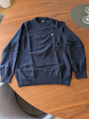 Mörkblå stickad tröja Lyle & Scott - Mörkblå stickad tröja från Lyle & Scott med rund halsringning och ribbade muddar. Tröjan har den klassiska gula logon broderad på bröstet och är tillverkad i mjukt material som passar perfekt till lager-på-lager-stil.