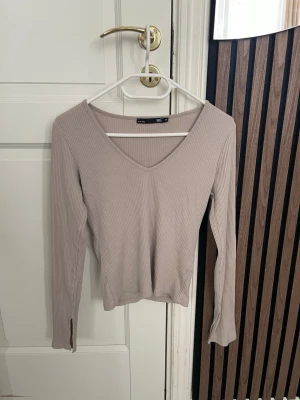Beige ribbad topp från 157 - Säljer en beige långärmad topp från 157 i ribbat material. Toppen har en djup V-ringning och smal passform, perfekt att styla med jeans eller kjol. Materialet är stretchigt och mjukt för extra komfort.