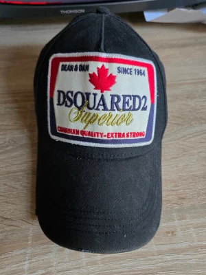 Svart DSQUARED2 keps med patch - Svart keps från DSQUARED2 med stor broderad patch framtill i rött, vitt och gult. Texten 'Superior' och ett rött lönnlöv ger kepsen en tydlig kanadensisk vibe. Justerbar baktill med broderad text 'FAMILY BUSINESS'. Klassisk böjd skärm och bomullsmaterial.