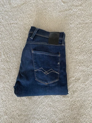 Mörkblå slim fit Replay anbass jeans - Snygga mörkblå slim fit jeans från Replay i modell anbass med klassisk femficksdesign och kontrastsömmar. Jeansen har en diskret broderad detalj på bakfickan och bälteshällor. Perfekta för en stilren och avslappnad look.