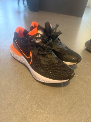 Svarta Nike löparskor  - Snygga svarta Nike löparskor med orange swoosh och detaljer. Ovandelen är i mesh för bra andningsförmåga och sulan är vit med en sportig look. Skorna har klassisk snörning och låg profil som passar till löpning och träning.
