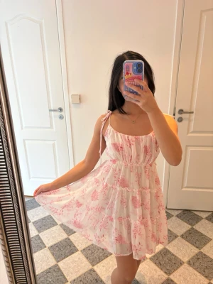Vit och rosa blommig sommarklänning - Kort sommarklänning med rosa blommigt mönster. Klänningen har flera lager så att inget syns igenom. Tunna axelband som man kan knyta om och justera. Klänningenär ca 80 cm lång från axelbanden. 