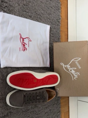 Gråa Christian Louboutin skor - Snygga gråa sneakers från Christian Louboutin med klassisk röd sula och vita kanter. Skorna har en stilren design i mocka och matchande gråa skosnören. Perfekta för dig som vill ha något exklusivt och trendigt i garderoben. Storlek 42 helt nya