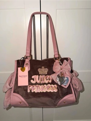 Juicy Couture rosa handväska - Superfin rosa och brun handväska från Juicy Couture med broderad text 'Juicy Princess', gulddetaljer och en stor rosett. Väskan har dubbla handtag, innerfoder med logga och ett hjärtformat hänge. Materialet är mjuk sammet och detaljer i skinnimitation.