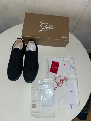 Svarta Louboutin sneakers med röd sula - Snygga svarta sneakers från Christian Louboutin med ikonisk röd sula och diskret logga på sidan. Skorna har en klassisk låg profil, svarta skosnören och ovandel i mocka. Perfekta för dig som vill sticka ut med exklusiv stil.