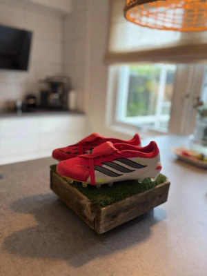 Röda Adidas fotbollsskor med dobbar - Snygga fotbollsskor från Adidas i rött med svarta detaljer och vita sulor. Skorna har klassiska dobbar för bra grepp på planen och är tillverkade i syntetmaterial. Perfekta för dig som vill sticka ut på fotbollsplanen.