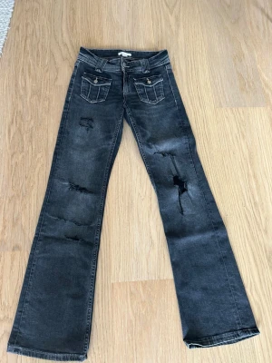 Grå slitna bootcut jeans Hina Tricot 158cm - Säljer ett par grå bootcut jeans med slitningar och trasiga detaljer på benen. Jeansen har två framfickor med lock och knapp, samt breda hällor för bälte. Från Gina Tricot 
