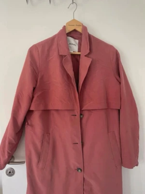 Rosa trenchcoat från Monki - Snygg rosa trenchcoat från Monki med klassisk krage, knäppning framtill och två sidofickor. Lång modell med avslappnad passform och stilren design. Perfekt för dig som vill ha en trendig och färgglad jacka till vår och höst. fläck och trasig skärphälla(se bilder)