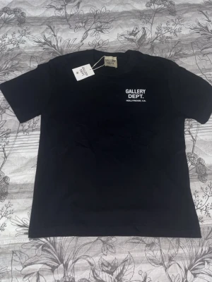 Svart Gallery Dept. t-shirt - Snygg svart t-shirt från Gallery Dept. med vit text på bröstet och ryggen där det står 'GALLERY DEPT. HOLLYWOOD, CA.'. Klassisk passform och rund hals. Perfekt för dig som gillar streetwear och enkel stil.