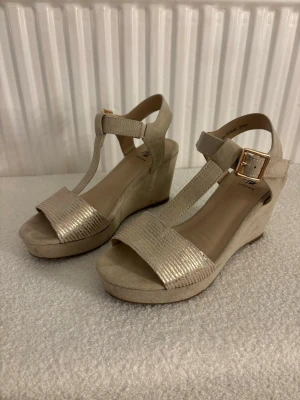 Beige kilklackade sandaler med spänne - Snygga beige sandaler med kilklack och T-rem. Främre remmen har ett diskret metallicmönster och sandalerna har ett guldfärgat spänne vid ankeln. Tillverkade i mockaimitation och syntetmaterial. Perfekta för dig som vill ha både höjd och komfort.