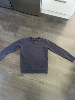 Mörkblå sweatshirt från Lyle & Scott - En stilren mörkblå sweatshirt med rund hals och lång ärm. Tröjan har en liten gul broderad logga på bröstet och ribbade muddar vid ärmslut och nederkant. Perfekt för en avslappnad look och enkel att matcha med jeans eller byxor.