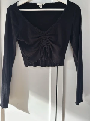 Svart croppad topp med snörning - Trendig svart croppad topp med lång ärm och v-ringning. Toppen har snygg snörning framtill som ger en rynkad effekt och är tillverkad i ett mjukt, ribbat material. Perfekt för dig som gillar en stilren men edgy look.