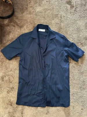 Mörkblå kortärmad skjorta från Zara - Snygg mörkblå skjorta från Zara i storlek M. Skjortan har korta ärmar, klassisk krage och knappar framtill. Tillverkad i ett mjukt och lätt material som passar perfekt till sommaren. Enkel och stilren design utan mönster.