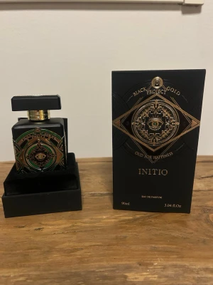 Initio Oud For Happiness parfym 90ml - Lyxig parfym från Initio, Oud For Happiness, i en svart flaska med gulddetaljer och gröna inslag. Flaskan har en fyrkantig form och ett dekorativt mönster med ett öga i centrum. Kommer i en matchande svart och guld ask. Innehåll: 90 ml Eau de Parfum.