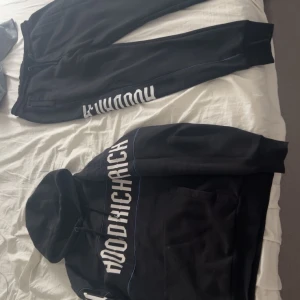 Svart hoodie från Hoodrich med tryck - Svart hoodie från Hoodrich med stor vit logga tryckt över bröstet och ärmarna. Tröjan har huva med snörning, långa ärmar och diskreta blå linjer som detalj. Perfekt för en streetwear-look och riktigt skön att bära.