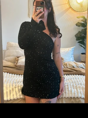 Svart glittrig oneshoulder klänning - Snygg svart kortklänning med glittriga paljetter och oneshoulder-design. Klänningen har lång ärm på ena sidan och är figurnära med en modern look. Perfekt för dig som vill sticka ut med en trendig och elegant stil.