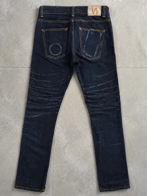 Nudie Jeans 31/32 - Snygga mörkblå jeans med skön passform och kontrastsömmar. Jeansen har klassiska fem fickor och unika slitningar på både fram- och baksida. Bakfickorna har coola detaljer och en patch i midjan. Perfekta för en trendig och avslappnad stil.
