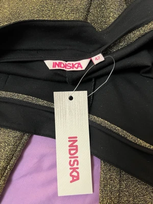 Svarta och guldglittriga mjukisbyxor Indiska - Snygga mjukisbyxor från Indiska i svart tyg med guldglittriga detaljer. Byxorna har en smal passform och resår i midjan för extra komfort. Perfekta för dig som vill ha något både bekvämt och trendigt med lite extra bling.