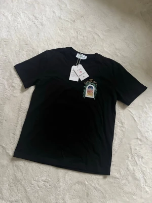 Svart t-shirt från Casablanca - Svart t-shirt från Casablanca med tryck på bröstet och stor färgglad grafik på ryggen med texten 'Avenida Casablanca' och ett fönster med arkitekturmotiv. Klassisk passform och rund hals. Perfekt för dig som vill sticka ut med en unik design.