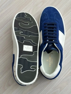 Blå och vita sneakers från Valentino - Snygga sneakers i blå mocka och vitt läder från Valentino. Skorna har svarta skosnören, cool sula med mönster och diskreta nitar på hälen. Perfekta för dig som vill sticka ut med stilrena detaljer och lyxig känsla.