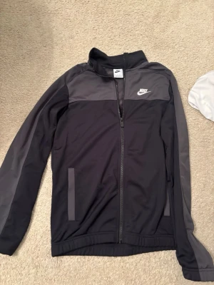 Svart vindjacka från Nike - Snygg svart vindjacka från Nike med grå detaljer på axlar och ärmar. Jackan har dragkedja framtill, hög krage och två fickor. Perfekt för dig som gillar sportig stil och vill ha något lätt och bekvämt.