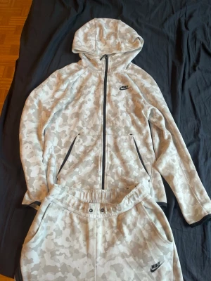 Nike camo set med hoodie och byxor - Nike set med hoodie och byxor i ljusgrått camomönster. Hoodien har dragkedja, fickor och svart Nike-logga på bröstet. Byxorna har resår i midjan, snörning och Nike-logga på benet. Mjukt material, perfekt för chill eller träning.