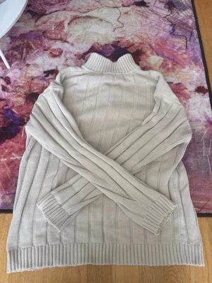 Beige stickad tröja med halv dragkedja GANT - Snygg beige stickad tröja med ståkrage och halv dragkedja framtill. Tröjan har ribbade muddar vid ärmslut och nederkant samt diskreta vertikala ränder i stickningen. Perfekt för lager på lager och enkel att matcha. GANT