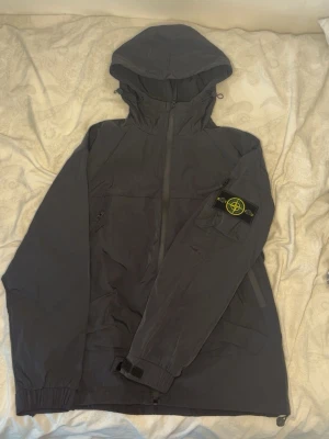 Stone Island vindjacka - Snygg svart vindjacka från Stone Island med ikonisk patch på ärmen. Jackan har huva, dragkedja framtill och justerbara muddar. Tillverkad i lätt, vindtätt material som passar perfekt för blåsiga dagar. Två sidofickor med dragkedja och en ficka på ärmen. Tyvärr är tagen i själva jackan klippt av tidigare ägare men fick med den tagen som satt på dragkedjan