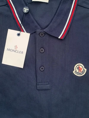 Marinblå pikétröja från Moncler - Snygg marinblå pikétröja från Moncler med klassisk krage och knäppning framtill. Kragen har röda och vita ränder och bröstet pryds av Monclers ikoniska logotyp. Perfekt för en stilren look.