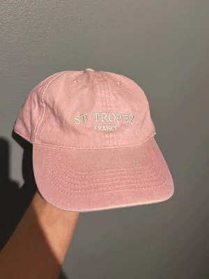 Rosa keps med St Tropez-brodyr - Säljer en ljusrosa keps med broderad text 'ST. TROPEZ FRANCE' framtill. Kepsen har böjd skärm, justerbart spänne bak och är tillverkad i bomullstyg. Perfekt accessoar för en chill och trendig look.