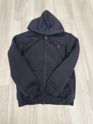 Svart hoodie från Polo Ralph Lauren REP - Säljer en svart hoodie från Polo Ralph Lauren med dragkedja och huva. Klassisk röd logga broderad på bröstet. Hoodien har fickor framtill och ribbade muddar vid ärmar och nederkant. Perfekt för en chill och stilren look.