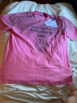 Rosa Acne Studios t-shirt med tryck - Snygg rosa t-shirt från Acne Studios med svart texttryck 'Acne Studios STOCKHOLM 1996' på bröstet. Klassisk passform med rund hals och korta ärmar. Perfekt för dig som gillar streetwear och vill sticka ut med en ikonisk look.