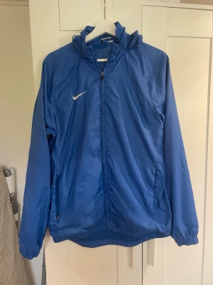 Blå vindjacka från Nike med huva - Snygg blå vindjacka från Nike med dragkedja framtill, huva och två sidofickor med dragkedja. Jackan har elastiska muddar vid ärmsluten och är tillverkad i ett lätt, vindtätt material. Perfekt för blåsiga dagar och sportiga outfits.
