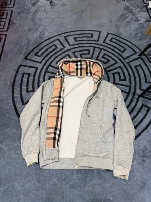  Burberry zipper  - Snygg grå zip-hoodie med klassiskt rutigt foder i huvan och dragkedja framtill. Hoodien har två fickor och ribbade muddar vid ärmslut och nederkant. Liten broderad logga på bröstet. Perfekt för en chill och stilren look.