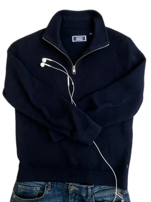 Mörkblå halvzip tröja Jack & Jones - Mörkblå stickad tröja från Jack & Jones med halvzip och hög krage. Tröjan har ribbade muddar vid ärmslut och nederkant, perfekt för lager på lager. Klassisk och enkel design som funkar till det mesta.