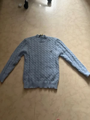Ljusblå kabelstickad tröja Polo Ralph Lauren - Snygg ljusblå kabelstickad tröja från Polo Ralph Lauren med rund halsringning och diskret logga broderad på bröstet. Tröjan har långa ärmar och ribbade muddar vid ärmslut och nederkant. Perfekt för dig som gillar klassisk och stilren look. Storlek M går att diskutera pris