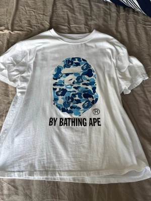 Vit BAPE t-shirt med blått camomönster - Säljer en vit t-shirt från By Bathing Ape med det klassiska BAPE-huvudet i blått camomönster på bröstet. T-shirten har korta ärmar och en relaxed passform. Perfekt för dig som gillar streetwear och vill sticka ut med en ikonisk design.