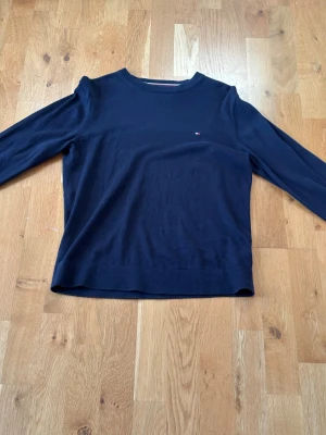 Mörkblå tröja Tommy Hilfiger - Klassisk mörkblå tröja från Tommy Hilfiger med rund halsringning och diskret logga på bröstet. Tröjan har långa ärmar och är tillverkad i ett mjukt, tunt material som passar perfekt till lager på lager. Enkel och stilren design.