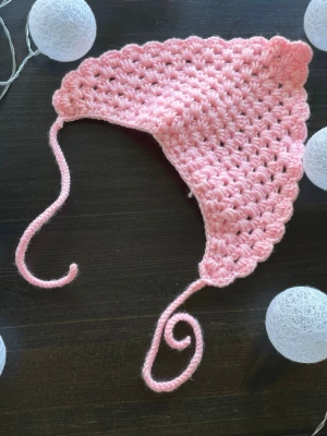 Rosa virkad mössa med snören - Söt rosa virkad mössa med snören att knyta under hakan. Mössan har ett vågigt kantmönster och är gjord i mjukt garn. Perfekt accessoar för dig som gillar en gullig och unik stil. Måtten på mössan är 24 cm bred 
