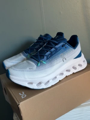Oncloud tilt blå vit  - Snygga sneakers från On i vitt och blått med mesh-ovandel och unika hål i den tjocka sulan. Skorna har blå snörning, diskreta loggor och detaljer. Perfekta för dig som gillar sportig och modern stil.