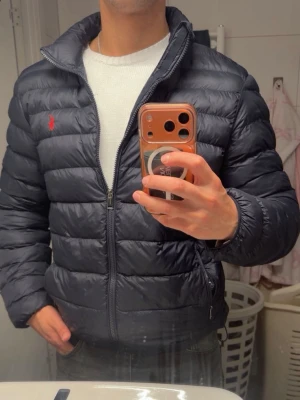 Ralph Lauren jacka  - Säljer en mörkblå pufferjacka från Polo Ralph Lauren med hög krage och dragkedja framtill. Jackan har det klassiska röda Polo-logot på bröstet och två sidofickor med dragkedja. Perfekt för kalla vinterdagar och riktigt snygg till jeans.