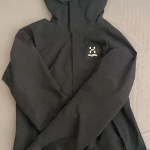 Svart vindjacka från Haglöfs - Säljer en svart vindjacka från Haglöfs med huva och dragkedja. Jackan är tillverkad i ett slitstarkt material och har en stilren design med logotypen broderad på bröstet. Perfekt för utomhusaktivitete