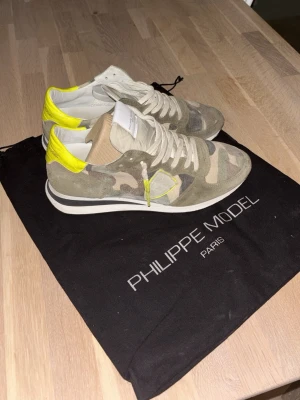 Camo sneakers från Philippe Model - Snygga sneakers från Philippe Model i beige och grönt camomönster med detaljer i neon-gult. Skorna har vita snören, vit sula och är tillverkade i en mix av mocka och textil. Perfekta för dig som vill sticka ut med en cool streetstyle. Box och dust bag får du med.