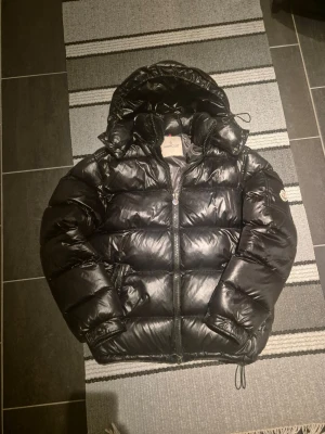 Svart dunjacka från Moncler - Säljer en svart, glansig dunjacka från Moncler med huva och dragkedja framtill. Jackan har en klassisk puffer-modell, dragsko i nederkant och Moncler-logga på ärmen. Insidan har ett serietidningsmönster. Perfekt för kalla vinterdagar. Jackan har ett litet håll på baksida armen men lagat o syns ej. Storlek 2 motsvarar ungefär S/M