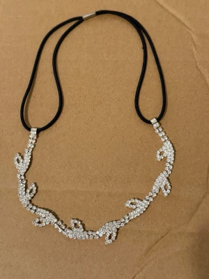 Glittrande hårband med strassdetaljer - Snyggt hårband med svart elastiskt band och gnistrande strass i ett bladliknande mönster framtill. Perfekt för att ge en extra touch till din frisyr. Materialet är elastiskt tyg och dekorationen är i silverfärgad metall med klara stenar.