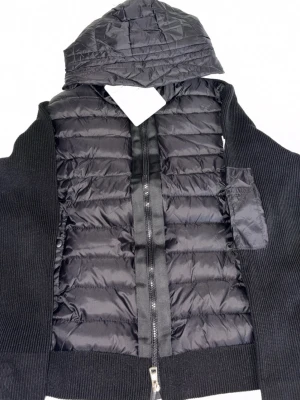 Svart Moncler cardigan - Snygg svart cardigan med quiltad framsida och stickade sidor samt rygg. Jackan har en praktisk huva och dragkedja framtill. En bröstficka med dragkedja ger extra detalj. Pris kan diskuteras