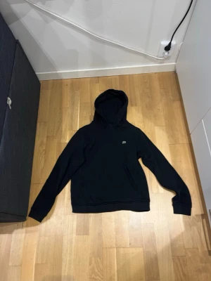 Svart hoodie från Lacoste  - En stilren svart hoodie med huva och lång ärm. På bröstet finns en liten diskret logga. Hoodien har klassisk passform och ribbade muddar vid ärmslut och nederkant. Perfekt för en avslappnad look.